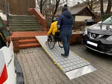 Frau schiebt Rollstuhlfahrer eine Rampe hoch.